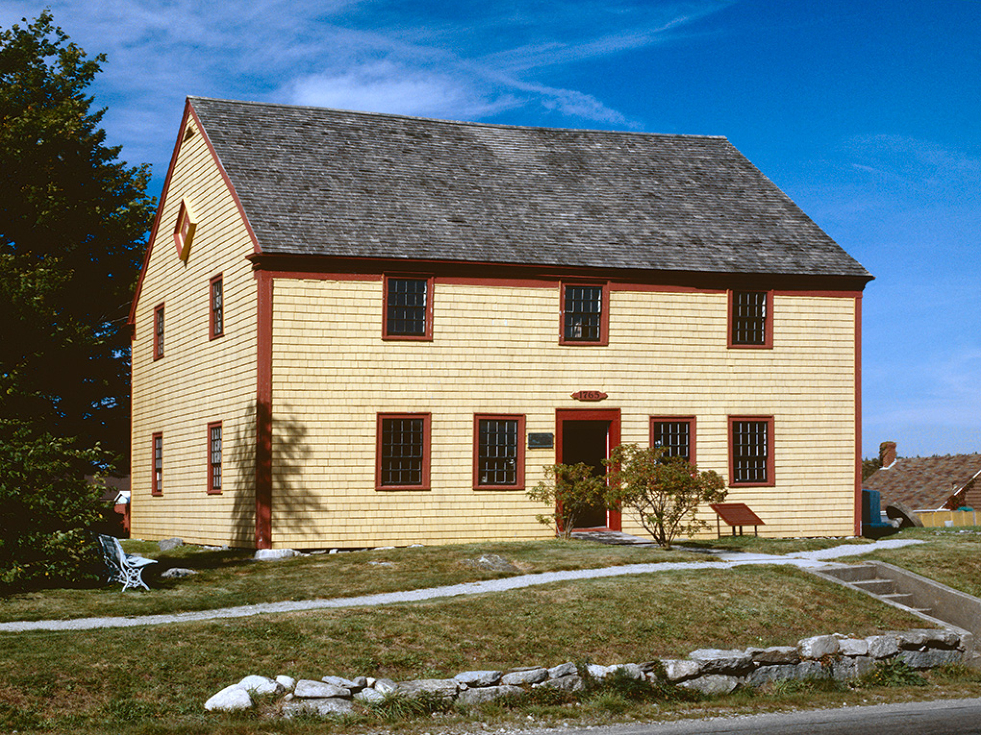 A two-story building with yellow shingle siding, a gray shingle roof, and red trim around the windows and doors. The building is situated on a grassy lawn with a stone wall in the foreground