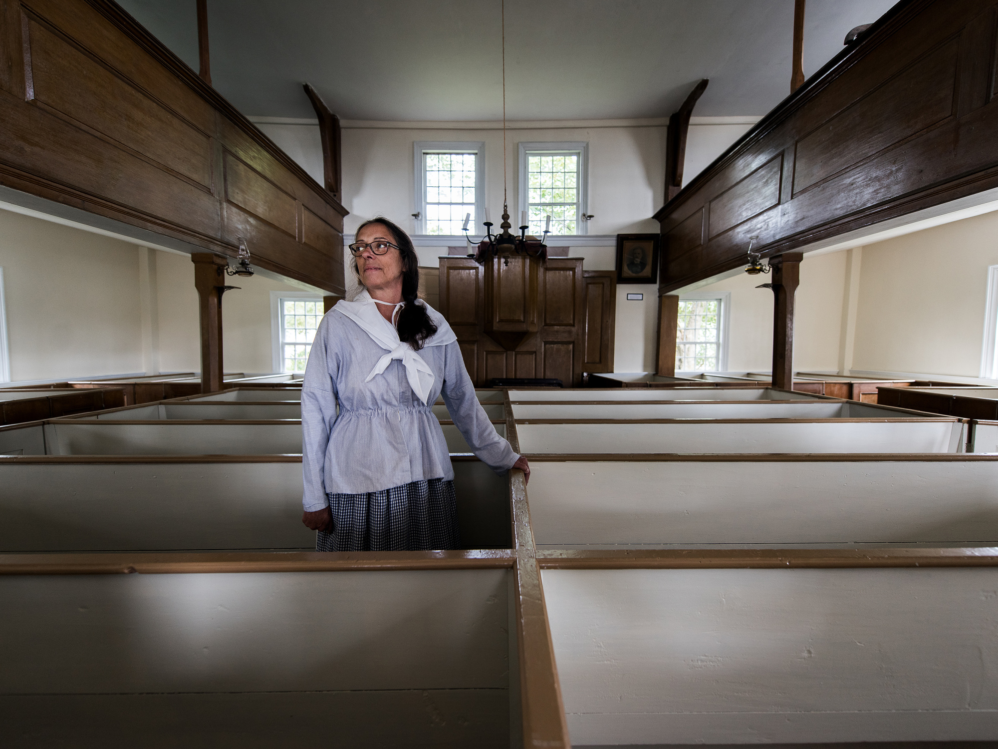 An interior view of a church. showing rows of empty pews, and a female costumed interpreter.