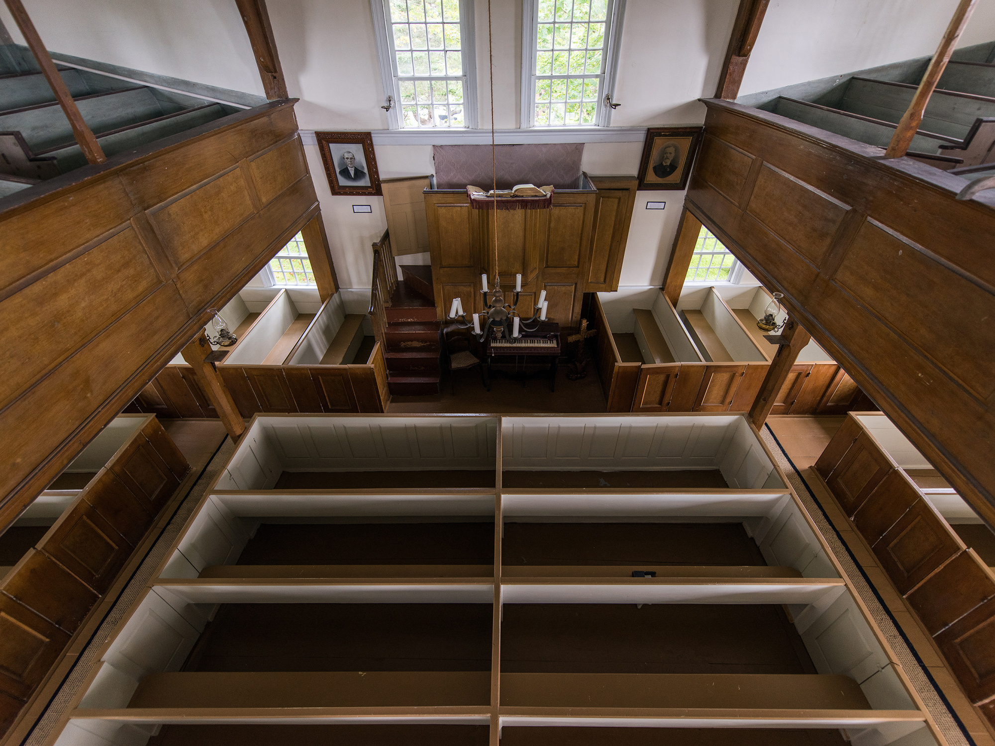 An overhead shot of a church interior, featuring rows of empty, light-colored pews with dark wooden trim. A raised pulpit with a book and several lit candles sits at the front, flanked by two framed portraits. Two tall windows are visible in the background. The church has dark wooden paneling on the walls