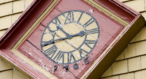 A close-up of a square, weathered clock face with Roman numerals and the year 1765, mounted on a building with yellow shingles.